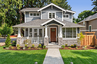 A two-story craftsman-style home with gray siding, white trim, and a stone-clad foundation features a stone walkway leading across a green lawn to a wooden front door