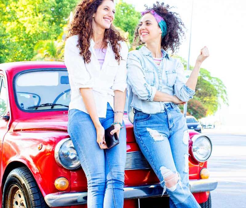 Two cheerful women with curly hair lean against the hood of a vintage red car, smiling warmly at each other - www.imperialnebraskainsurance.com